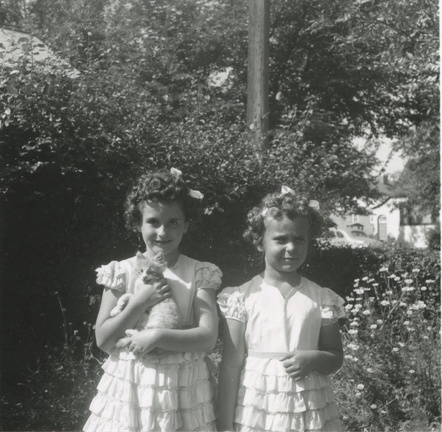Carole holding a kitten with Lynn in dresses near pine trees 1948?